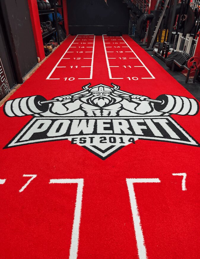 Ruohomatto-merkinnoilla-powerfit Red gym turf with white yard-line markings and a large PowerFIT logo featuring a muscular Viking lifting barbells; est. 2019.